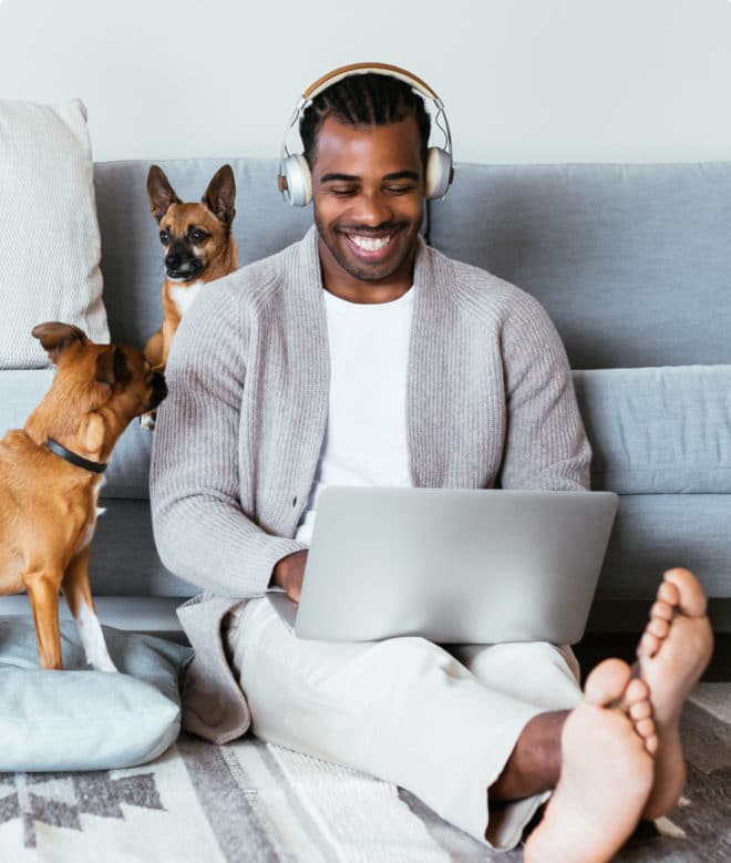 African American man looking at computer smiling, with two small dogs playing next to him on the couch